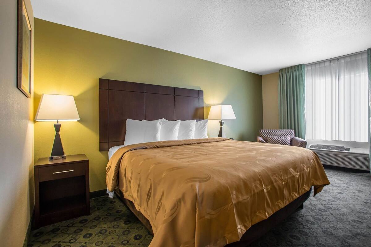 A hotel room with a large bed, two bedside lamps, a window with curtains, a chair, and a small table with a lamp.