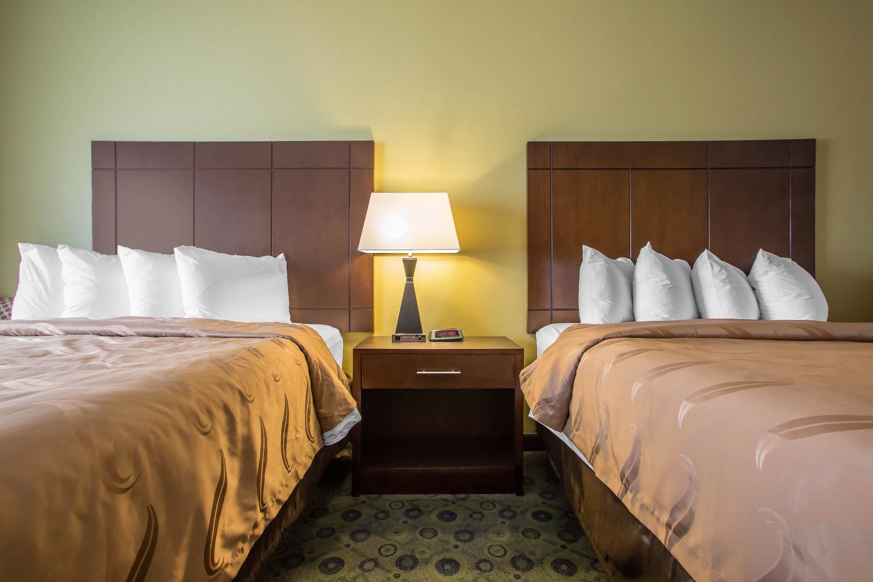 A hotel room with two beds, a nightstand with a lamp, and a digital clock.