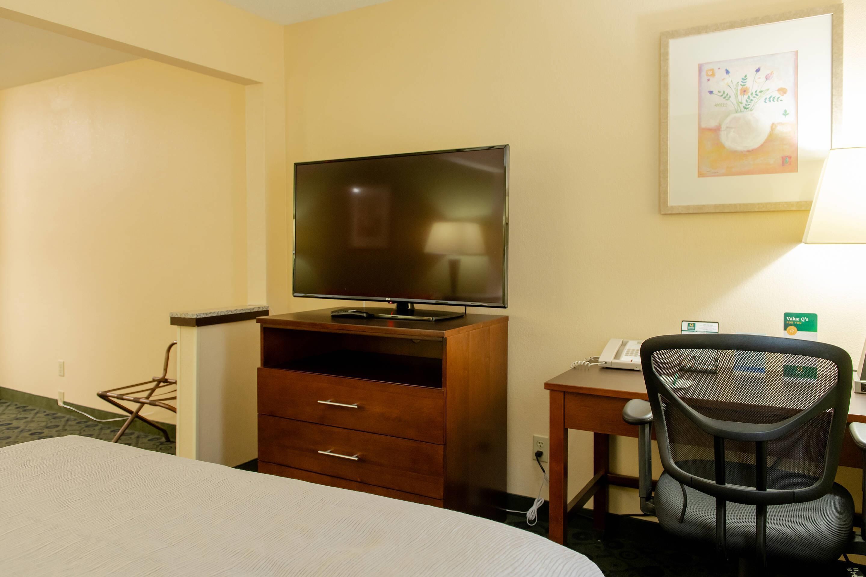 A hotel room with a bed, a desk with a computer, a TV on a wooden stand, and a chair.