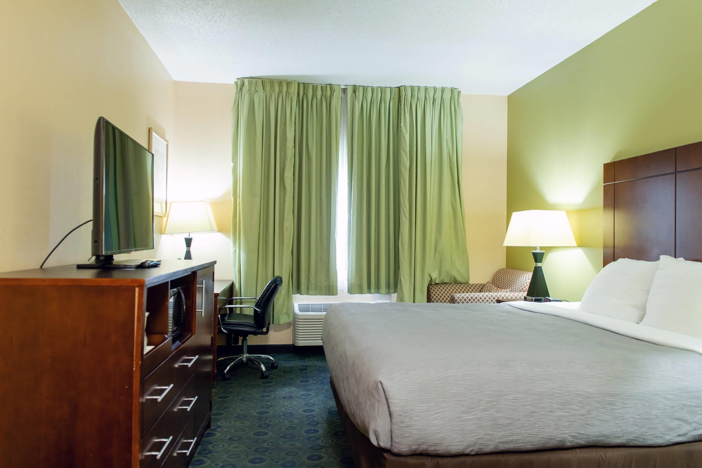 A hotel room with a bed, a desk with a TV, a chair, a lamp, and a window with curtains.