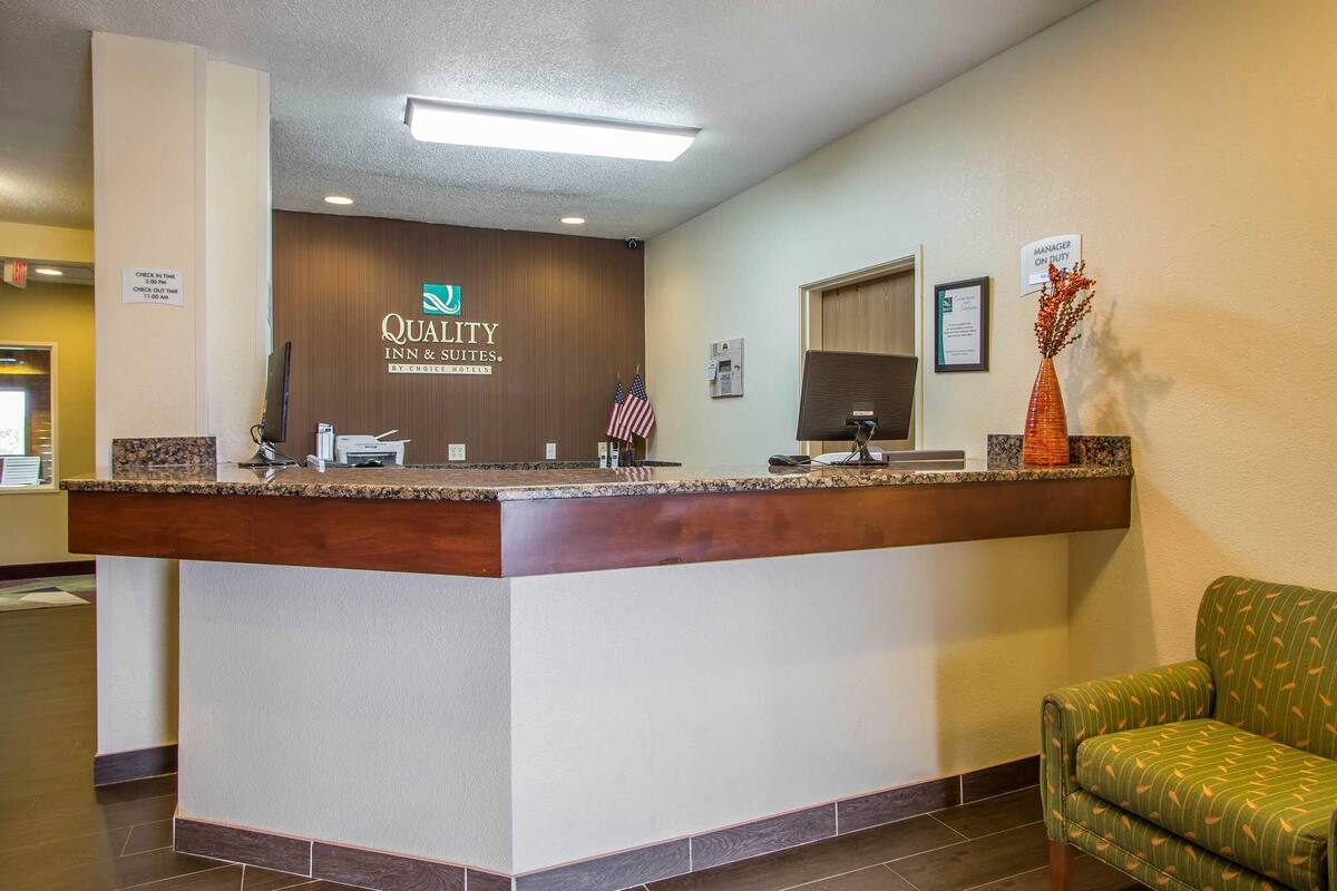 A hotel reception desk with a wooden counter, a sign reading 'Quality Inn & Suites by Choice Hotels,' an American flag, and a green patterned sofa.