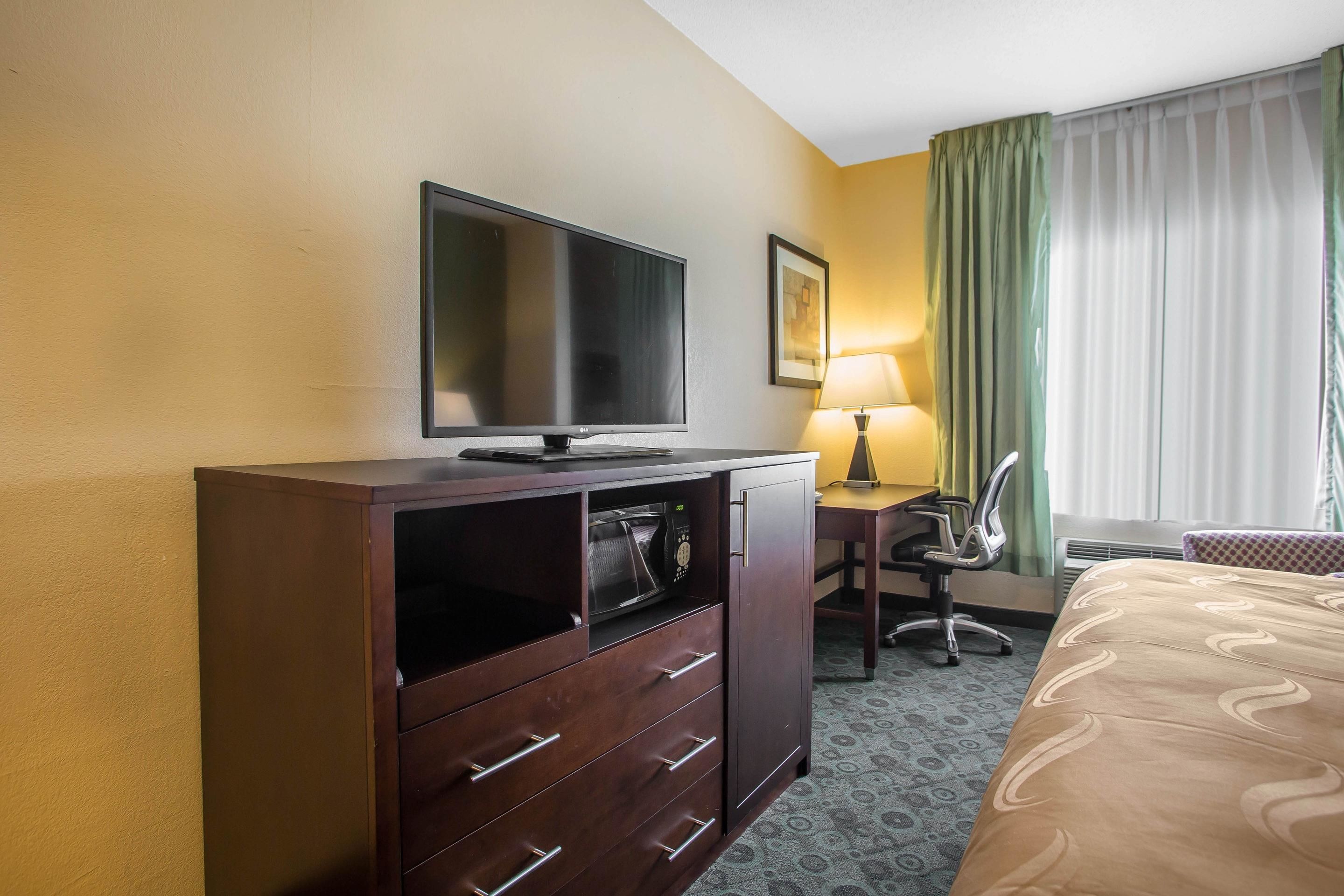 Habitación de hotel con cama, televisor en un mueble de madera, escritorio con silla y ventana con cortinas.
