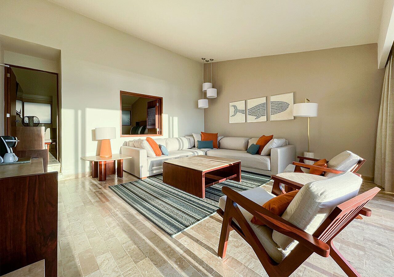 A modern living room with a sofa, coffee table, and armchairs. There is a wooden desk and a lamp.