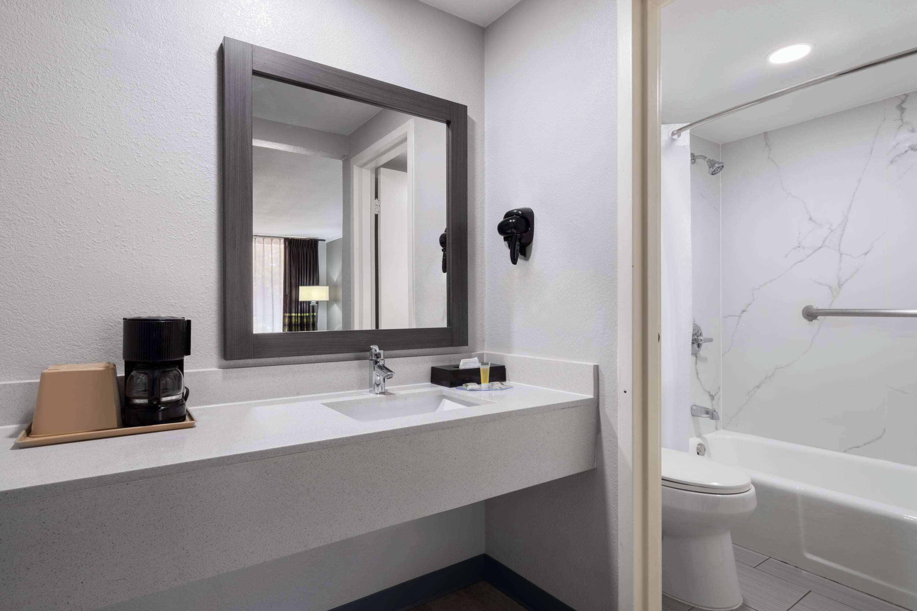 A modern bathroom with a white sink, a large mirror, a black hairdryer, and a white bathtub.