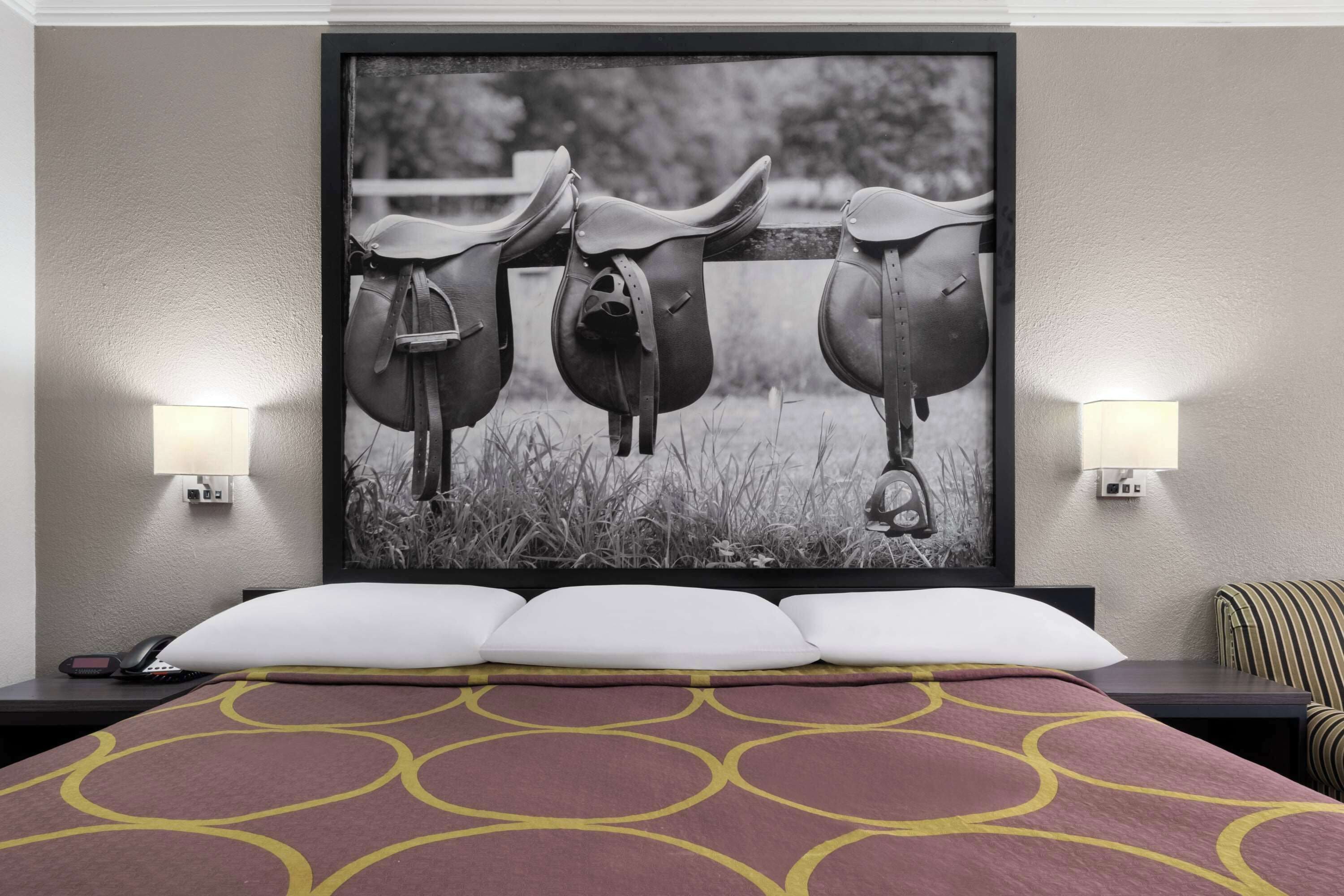 A hotel room with a bed, a framed picture of horse saddles, two wall-mounted lamps, and a striped chair.