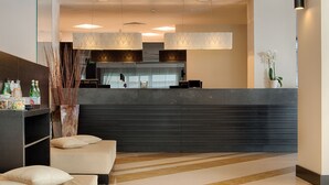 A modern reception area with a dark reception desk, a seating area with cushions, and a decorative vase with plants. - NH Trieste (Trieste)