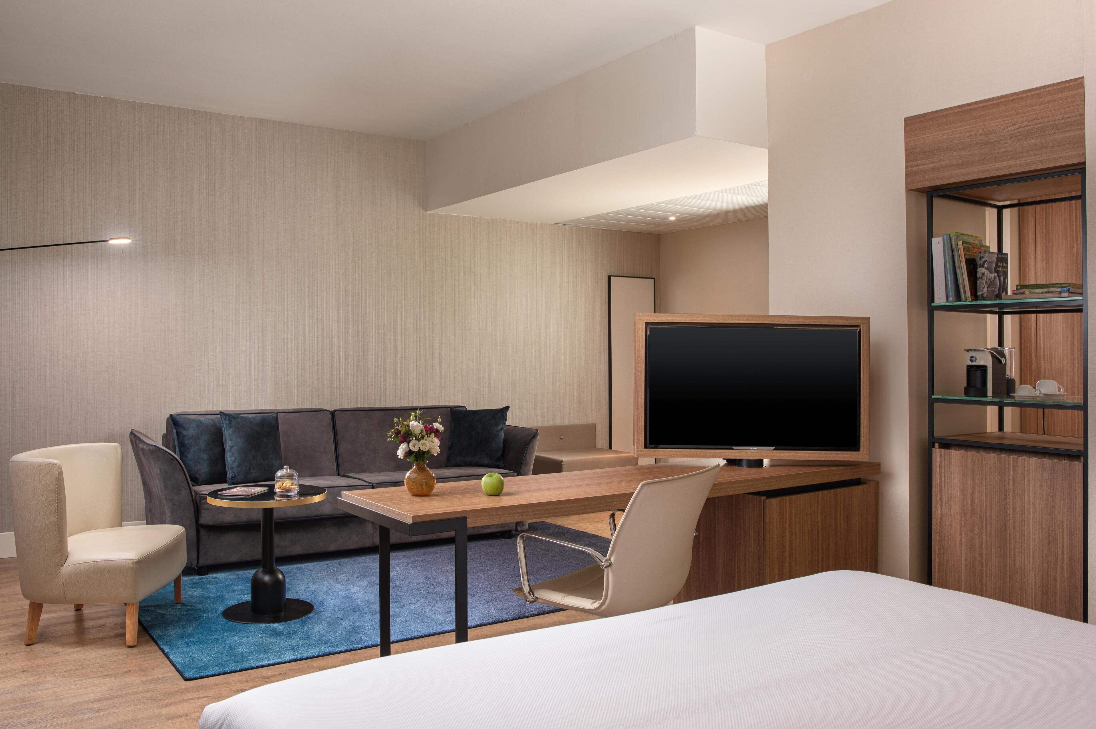 Una habitación de hotel moderna con sofá, una mesita con un florero y una manzana, televisión y estantería.