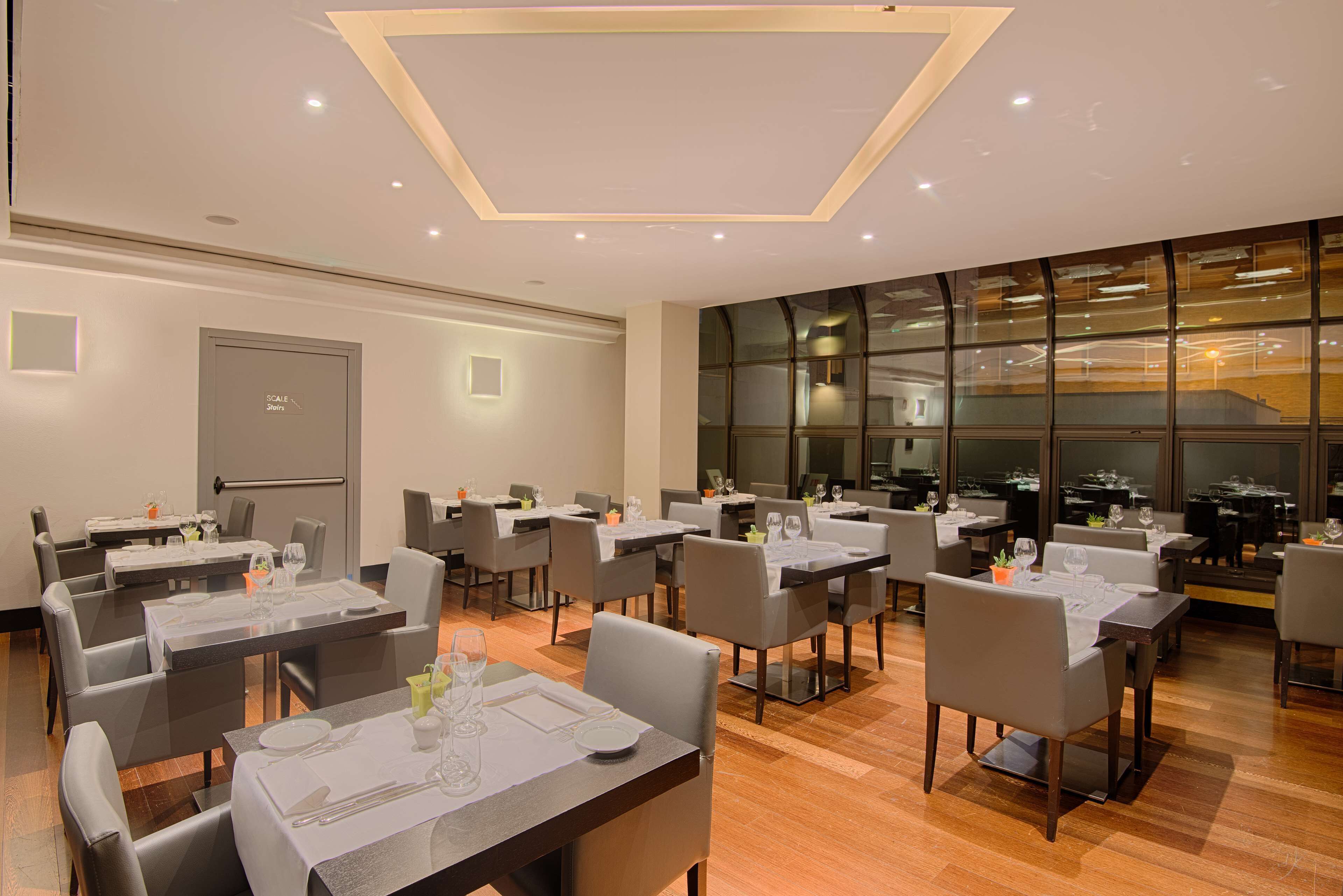 A modern dining area with multiple tables and chairs, a large window with a view, and a ceiling with recessed lighting.
