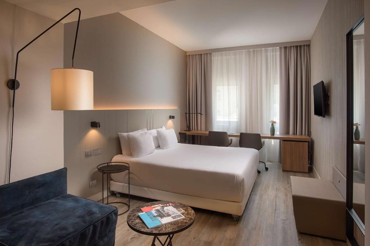 A hotel room with a bed, a desk, a chair, a sofa, a lamp, and a TV.