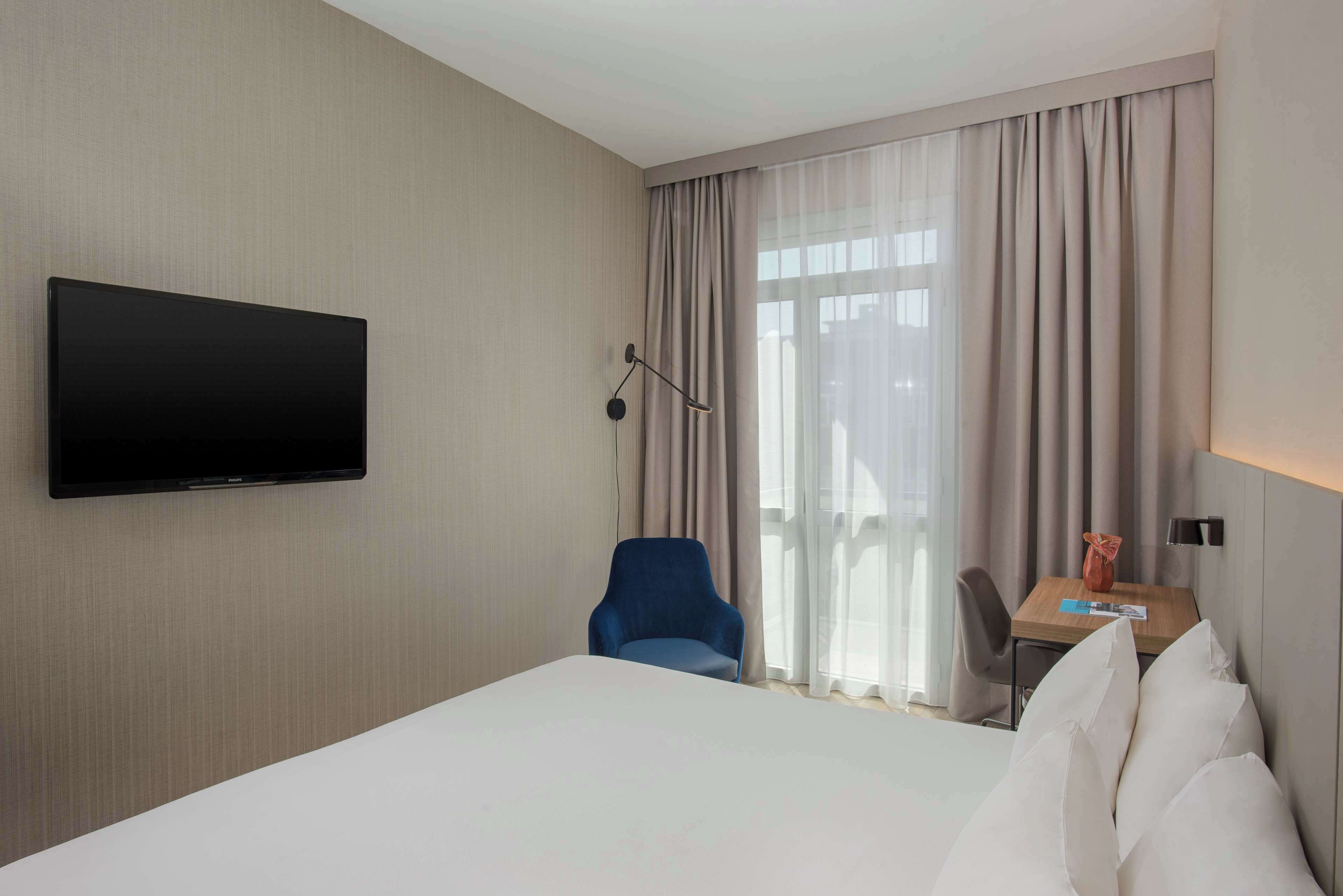 Habitación de hotel con una cama, un televisor de pantalla plana montado en la pared, un escritorio con una silla y una ventana con cortinas.