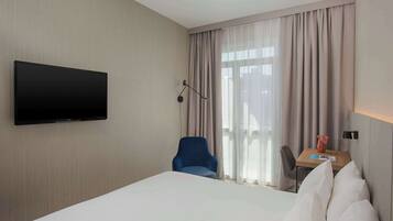 Ein Hotelzimmer mit einem Bett, einem an der Wand befestigten Flachbildfernseher, einem Schreibtisch mit Stuhl und einem mit Vorhängen versehenen Fenster.