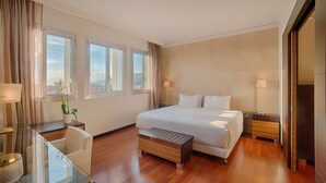 A hotel room with a large bed, a desk with a chair, a small table with a lamp, and a view of the city through the windows. - NH Trieste (Trieste)
