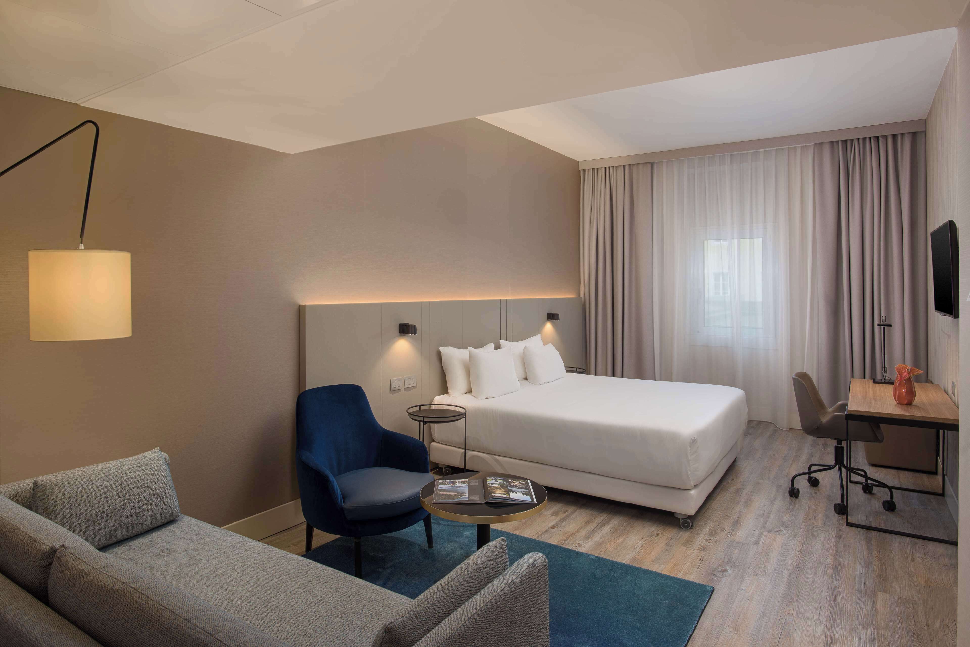 A hotel room with a bed, a desk, a chair, a sofa, and a lamp.