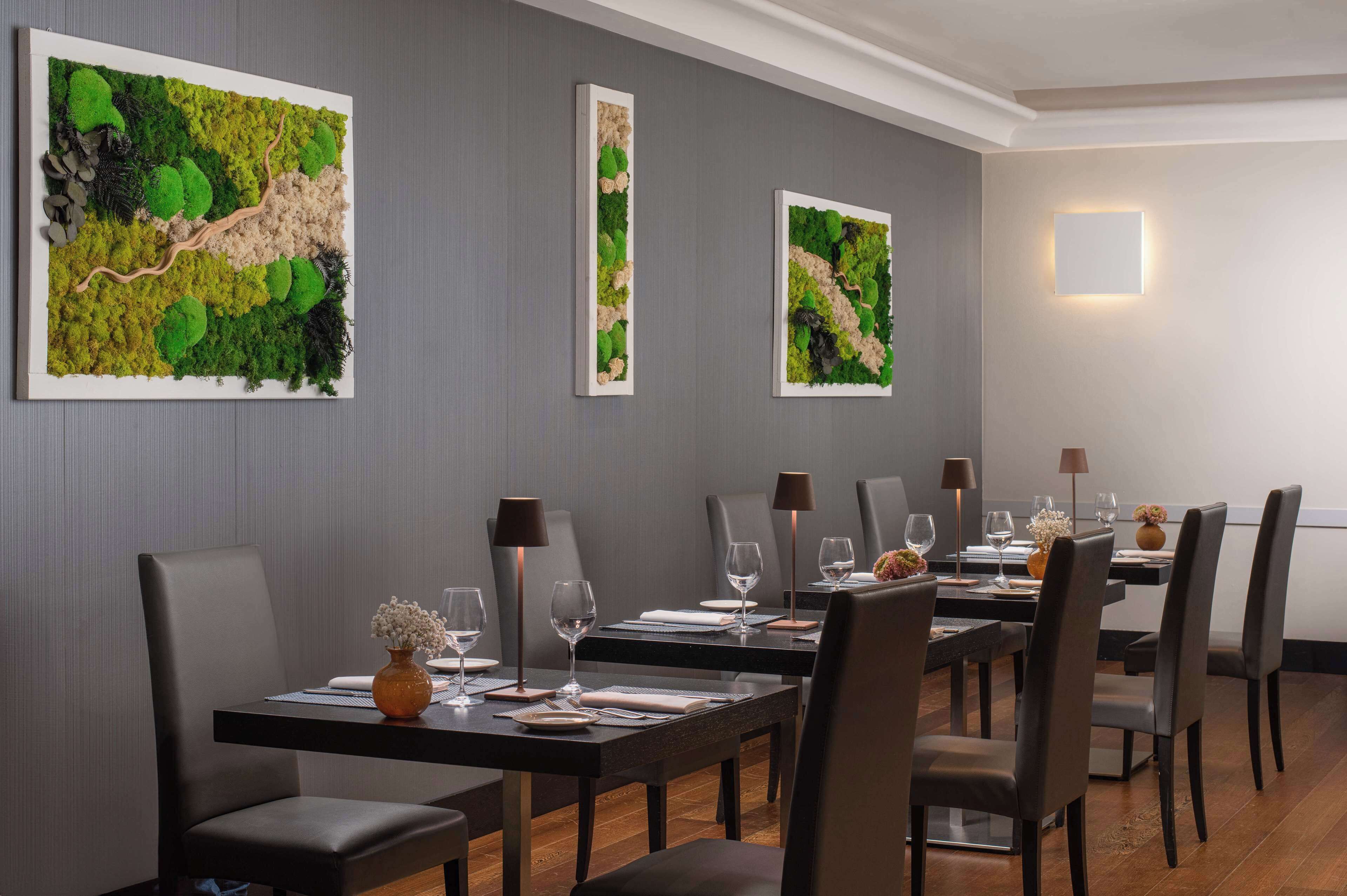 Un comedor con mesas dispuestas para seis comensales, cada una con un jarrón de flores, copas de vino y cubiertos. Las paredes están decoradas con grandes pinturas abstractas.