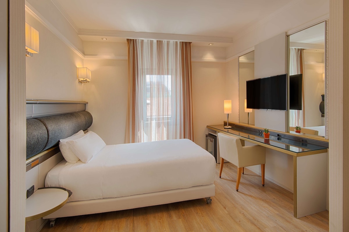 A hotel room with a bed, desk, chair, TV, and curtains.