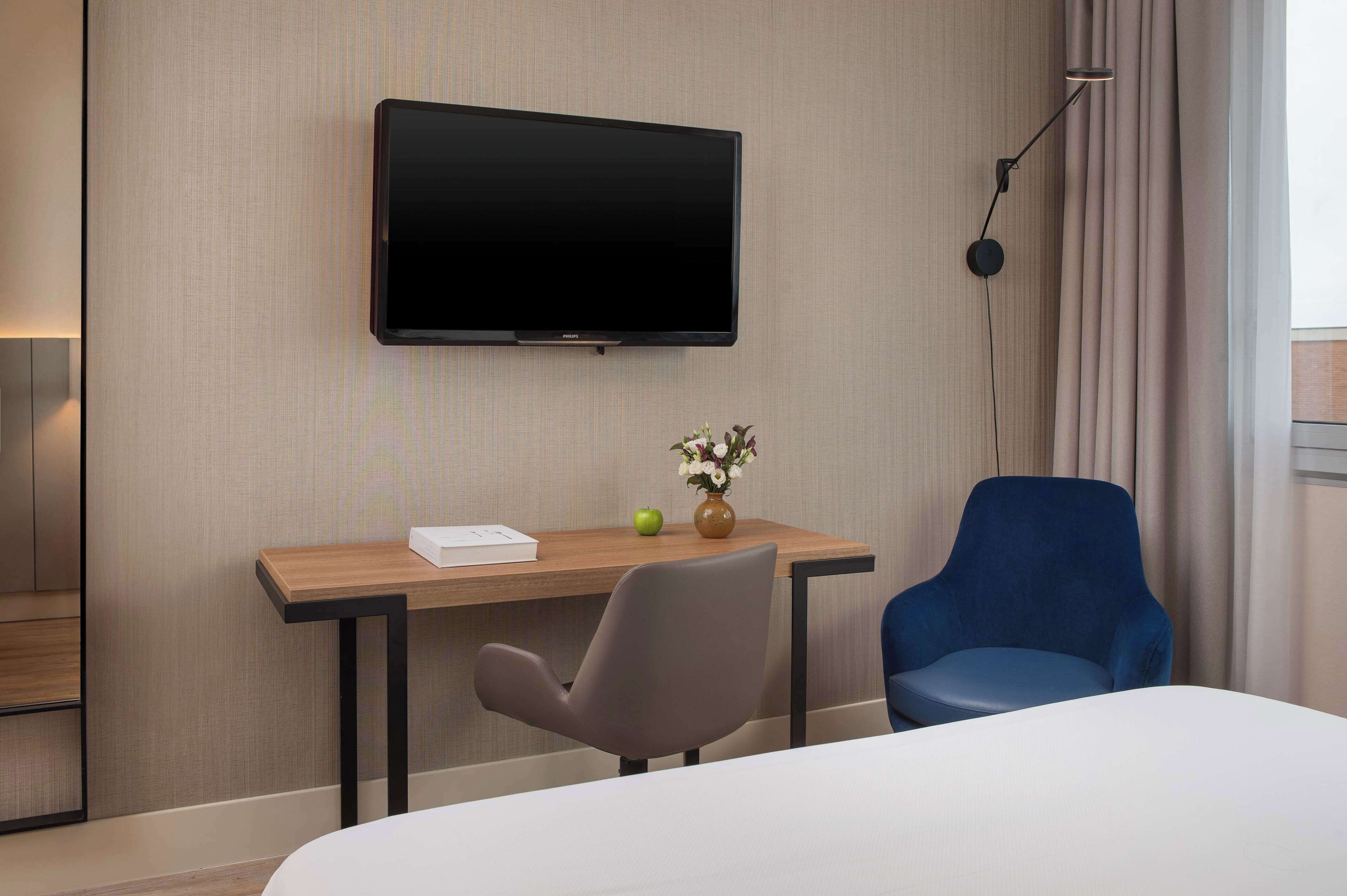 Une chambre d’hôtel dotée d’un téléviseur à écran plat fixé au mur, d’un bureau en bois agrémenté d’un vase de fleurs, d’un fauteuil de bureau gris, d’un fauteuil bleu et d’un lit avec une literie blanche.