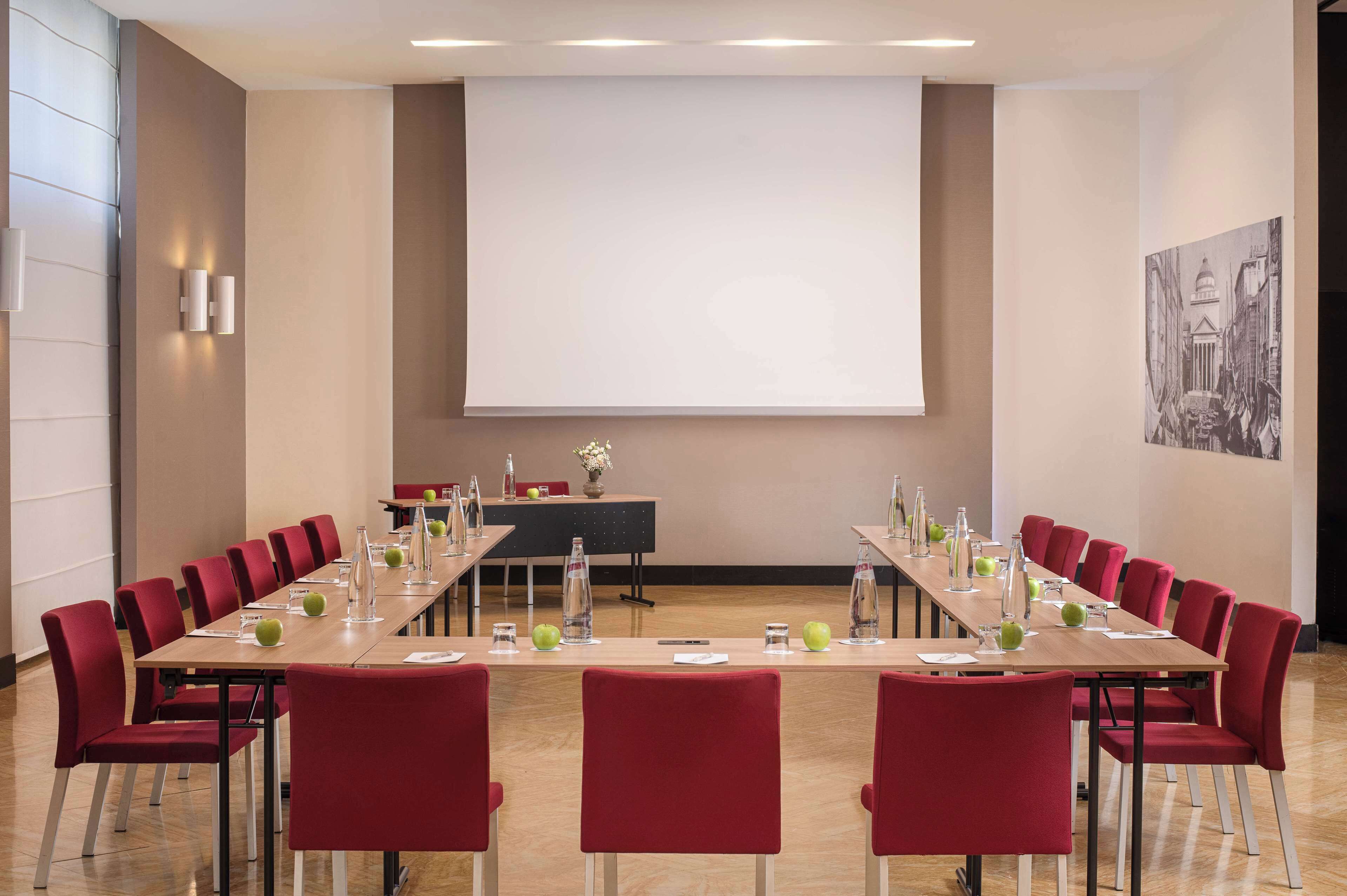 Un salón de conferencias con una larga mesa, sillas rojas, botellas de agua y una pantalla para proyector.
