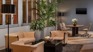 A modern living room with beige sofas, a glass coffee table, a large potted plant, a dining table with chairs, and a flat-screen TV mounted on the wall. - NH Trieste (Trieste)
