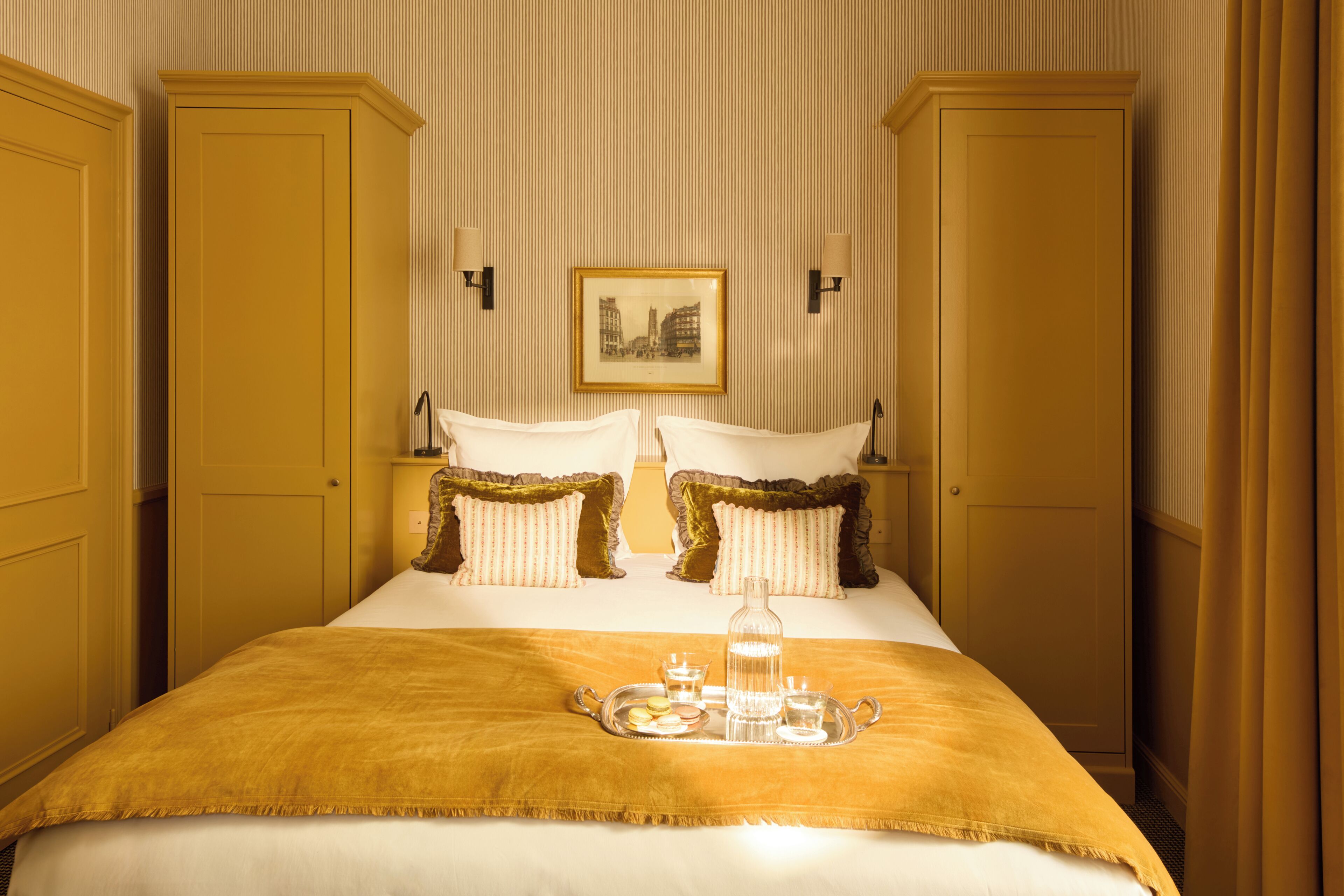 A hotel room with a bed, two nightstands, a framed picture on the wall, and a tray with glasses and a bottle on the bed.