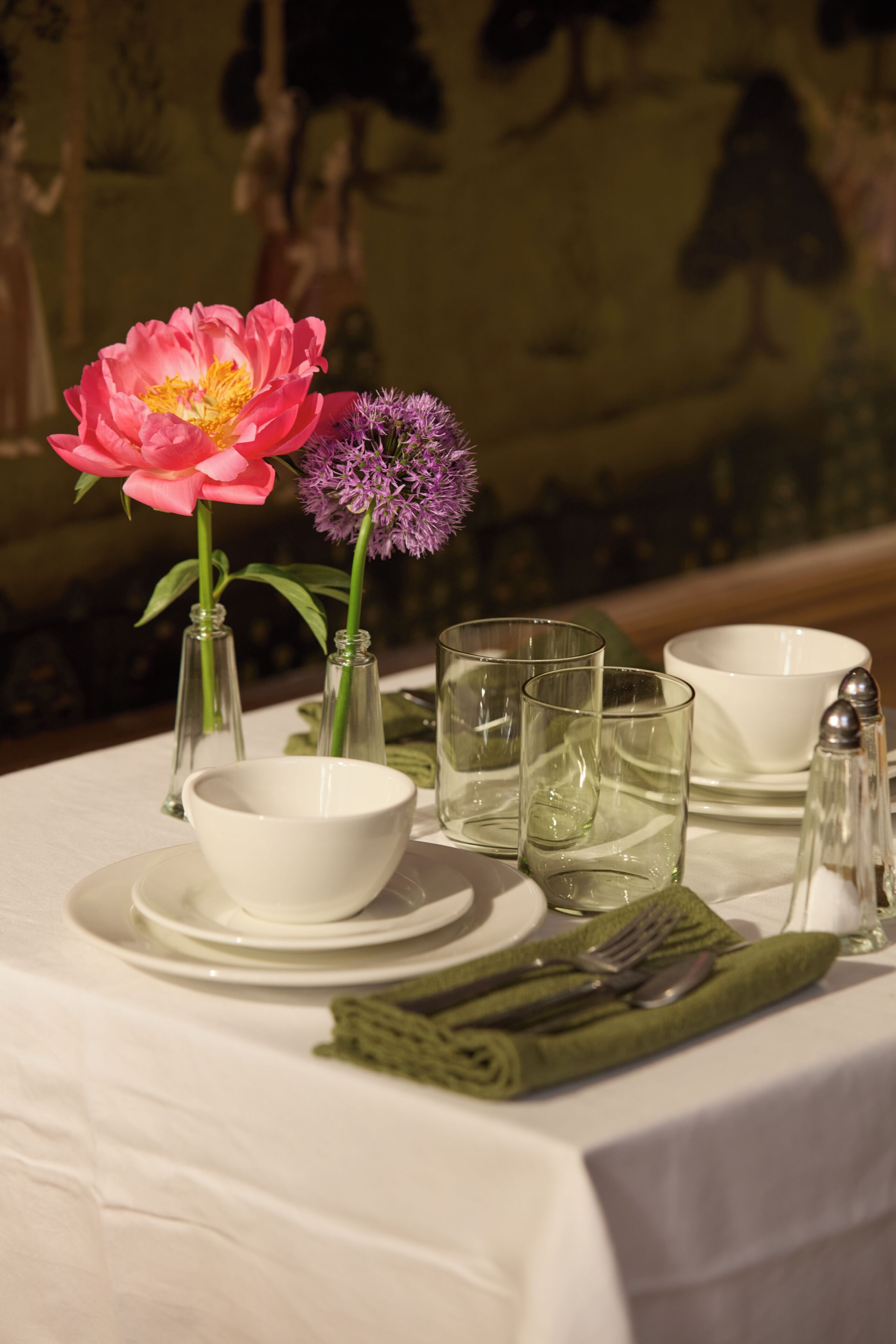 A table set with a white tablecloth, a pink flower, a purple flower, clear glasses, a white bowl, and a green napkin.