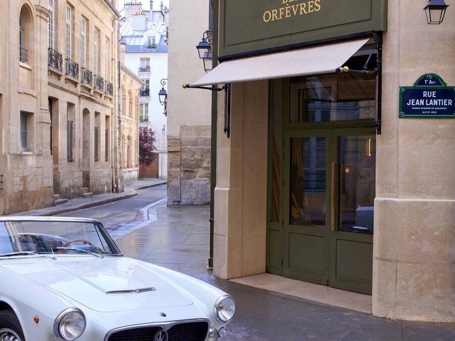 Un descapotable blanco clásico estacionado frente a un hotel llamado 'Hôtel des Orfèvres', con un cartel que indica 'RUE JEAN LANTIER'.