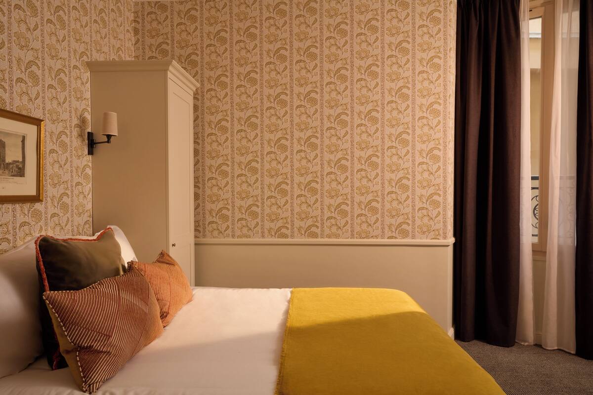 A hotel room with a bed, a nightstand, a lamp, a framed picture on the wall, and a window with curtains.