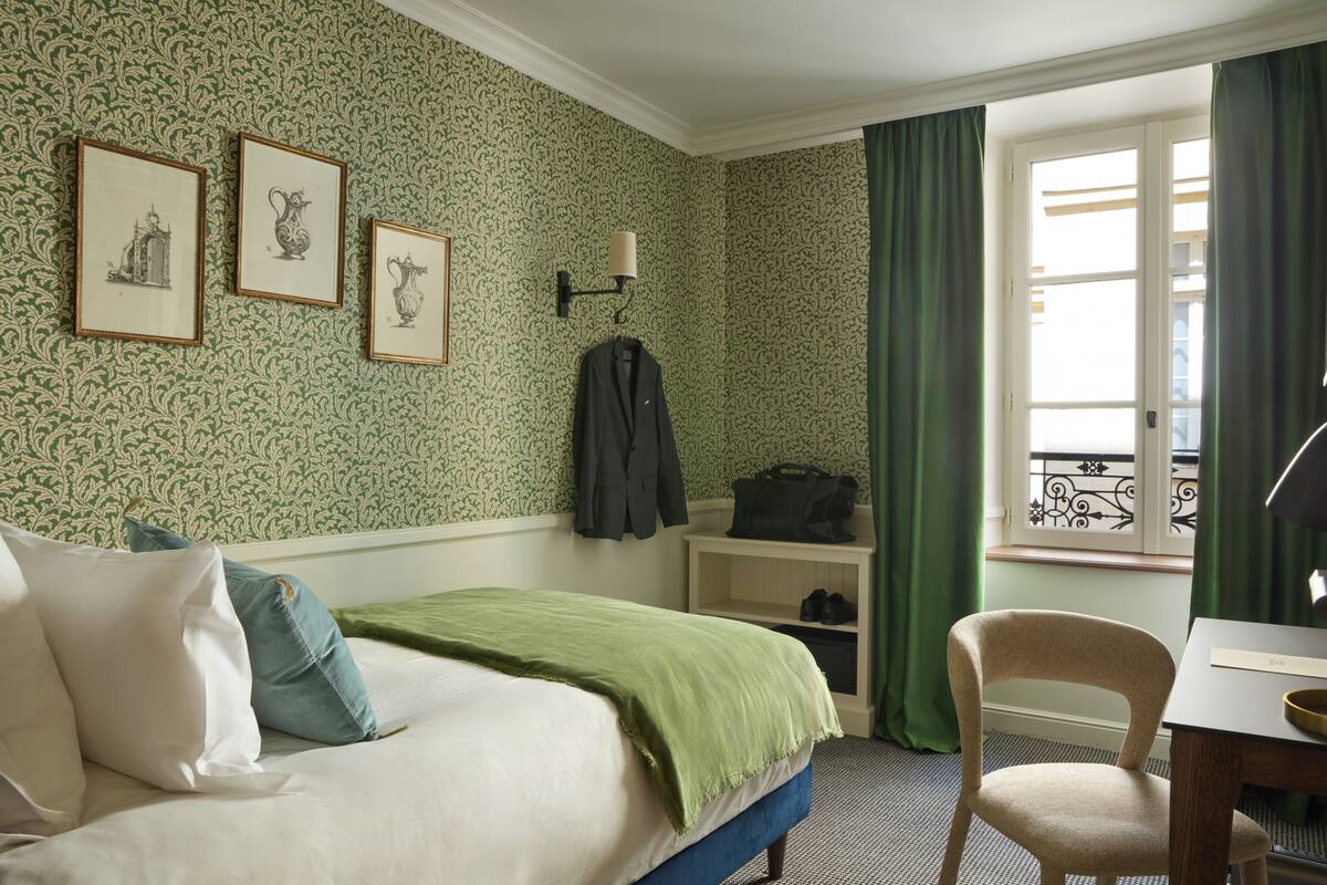 A hotel room with a bed, a chair, a desk, a coat rack, and a window with curtains.