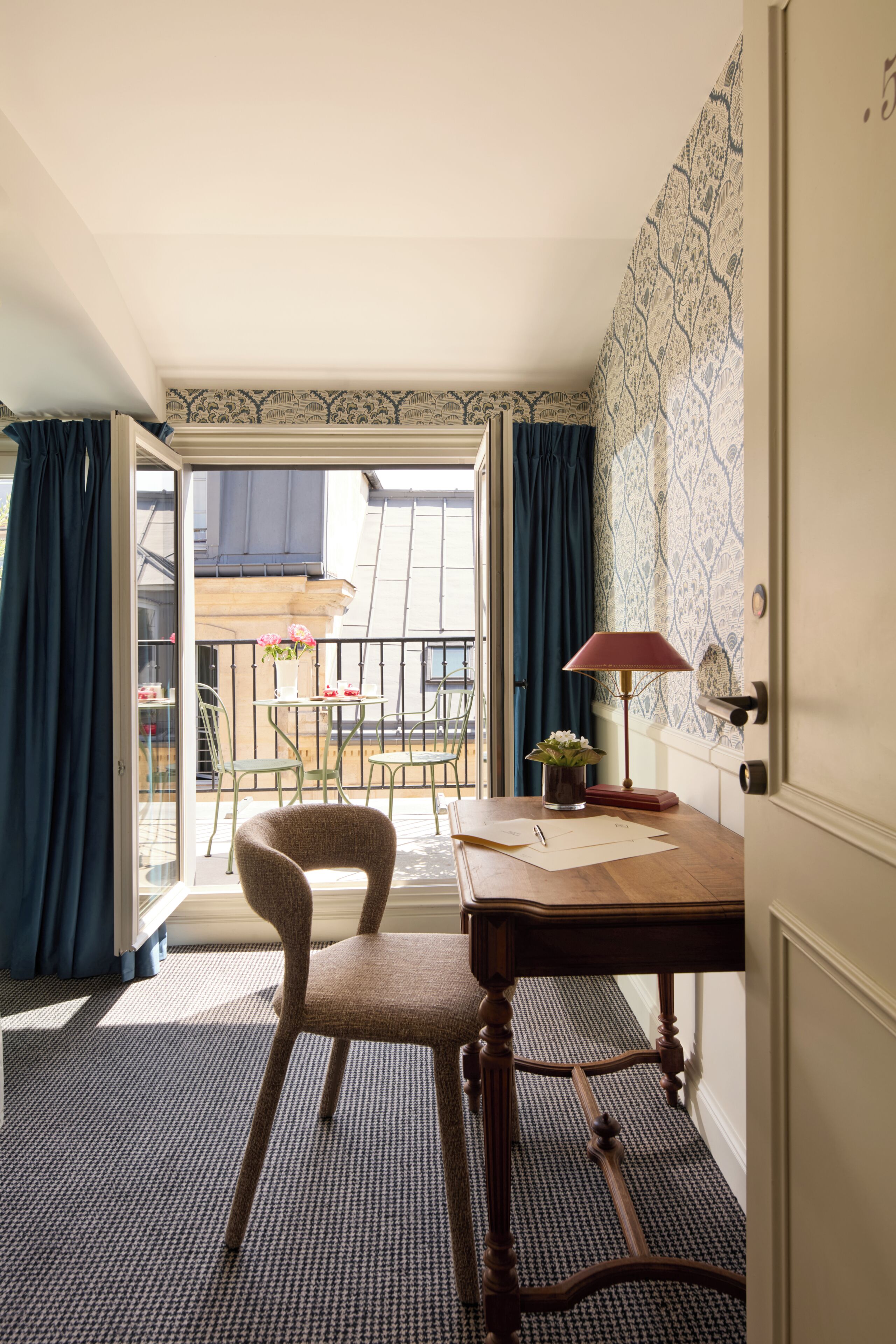 Una habitación con un escritorio de madera, una silla, una lámpara y vistas a un balcón exterior con una mesa y sillas.