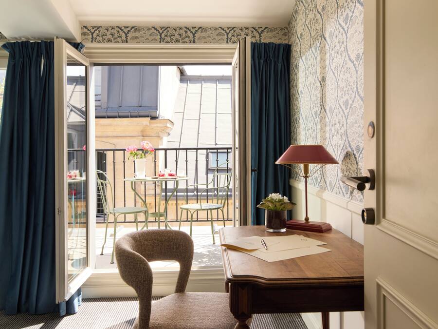 Una habitación con un escritorio de madera, una silla, una lámpara y vistas a un balcón exterior con una mesa y sillas.