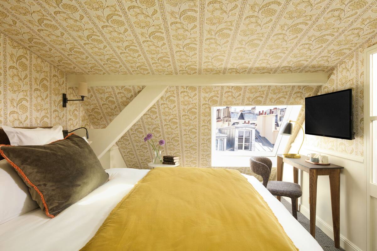 A bedroom with a bed, a desk, a chair, a TV, and a window with a view of buildings.