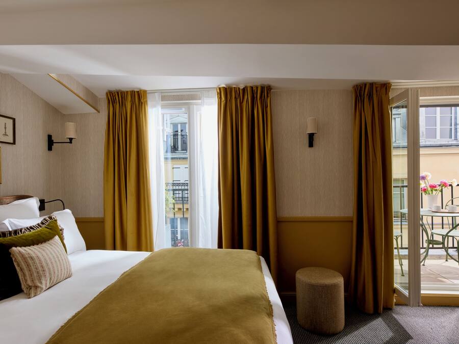Habitación de hotel con cama, un taburete pequeño, un balcón con mesa y sillas, y vistas al exterior.