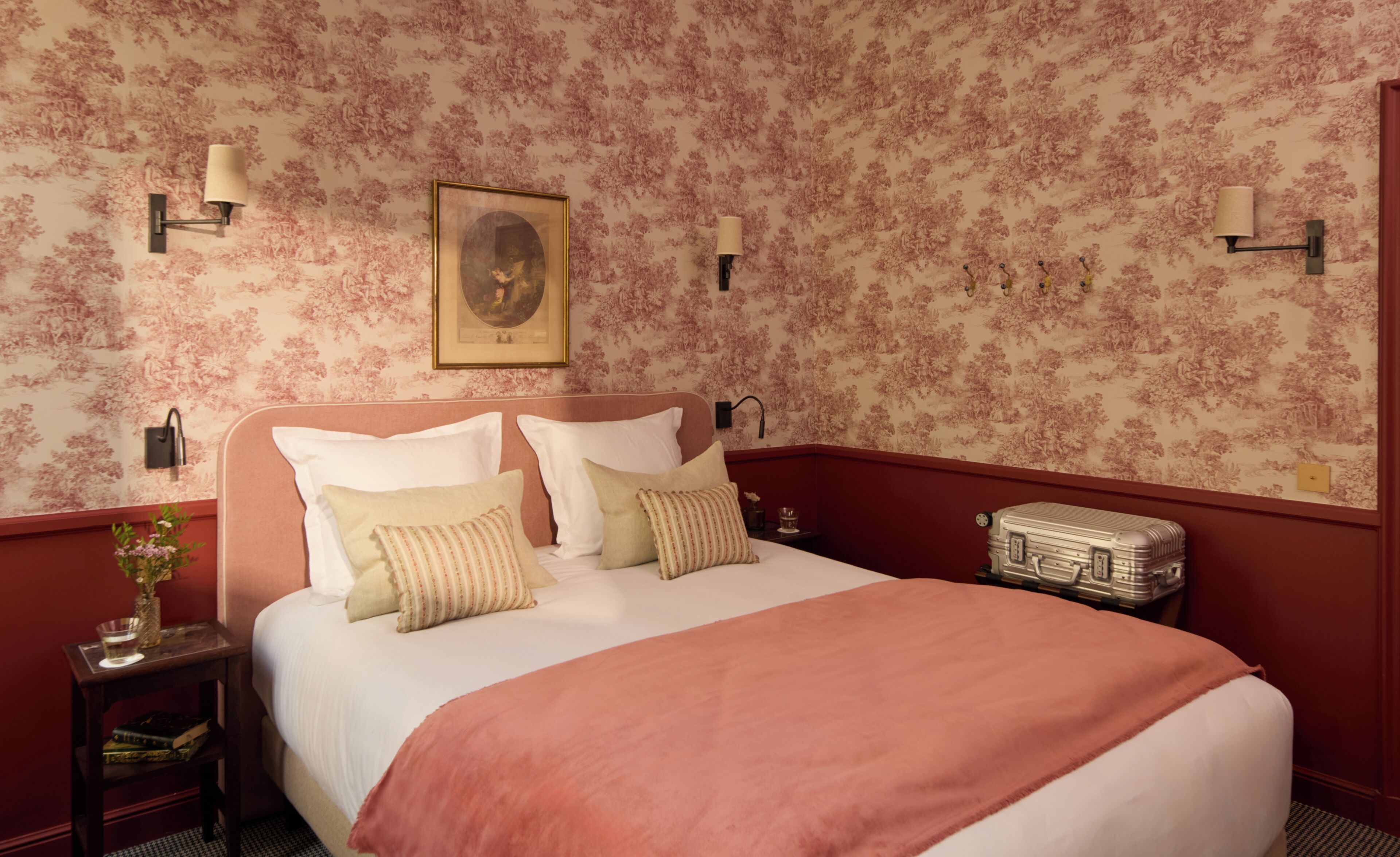 A bedroom with a floral wallpaper, a bed with white linens, two wall-mounted lamps, a framed portrait, and a vintage suitcase.