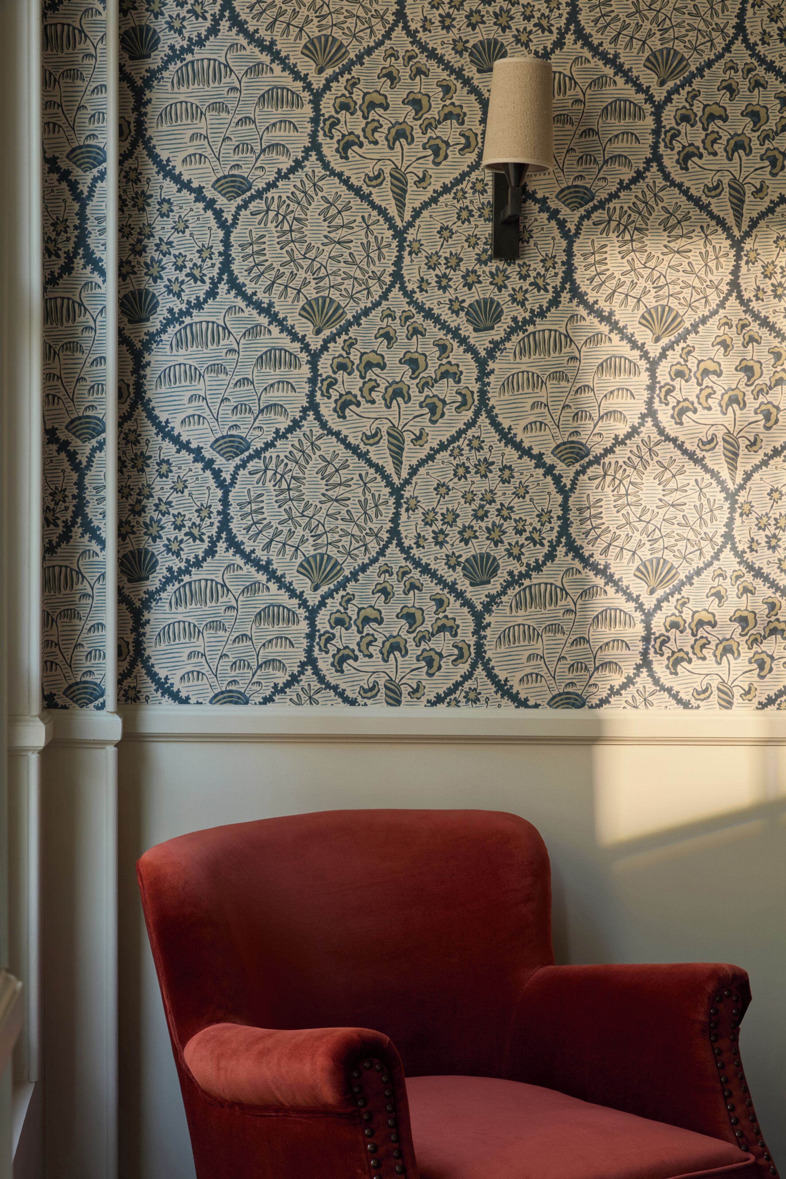 Quarto com papel de parede estampado, uma cadeira estofada vermelha e uma luminária fixada na parede.
