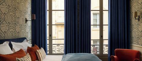 A hotel room with a bed, pillows, a chair, and a view through a window with curtains.