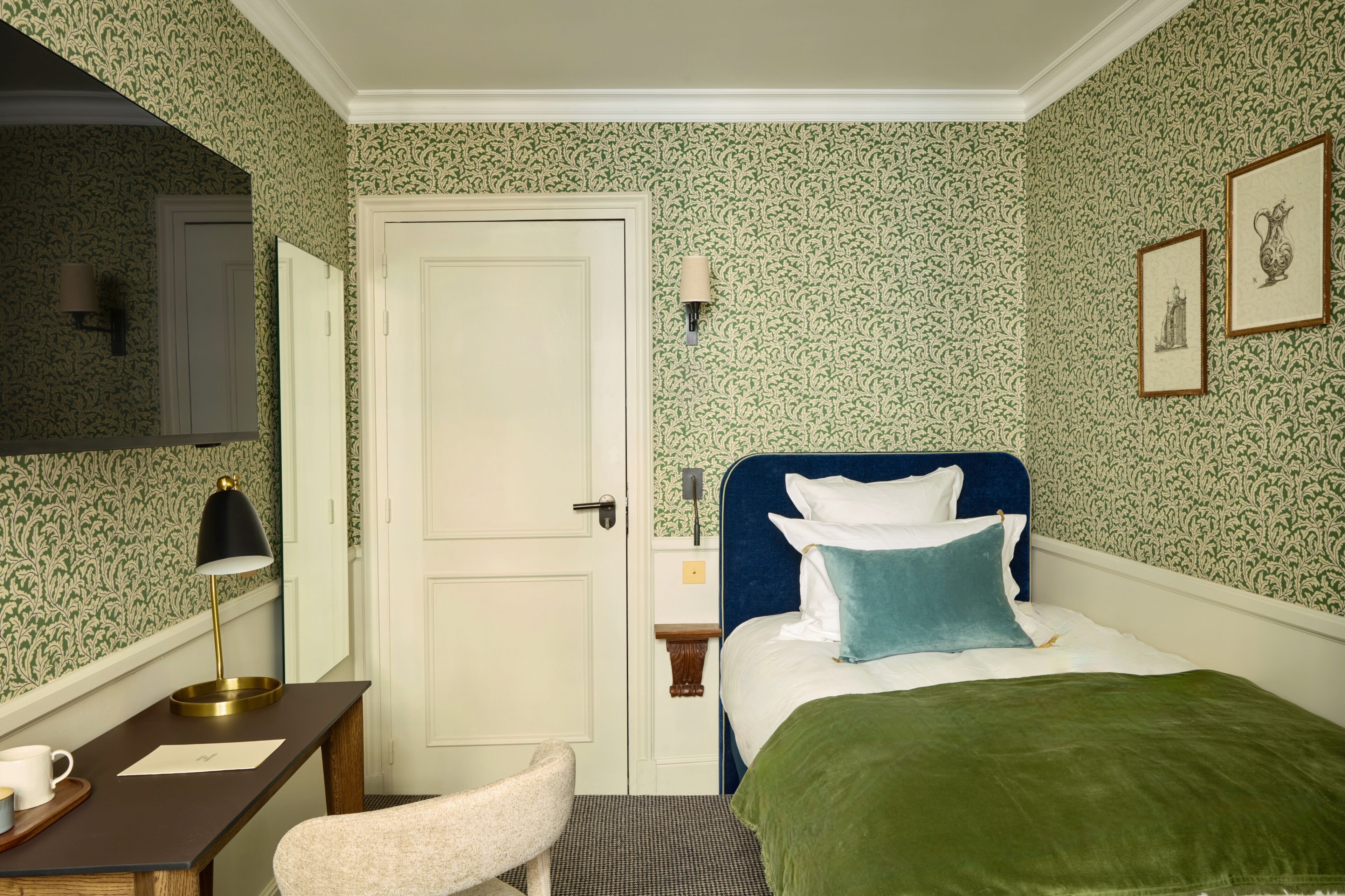 A hotel room with a bed, a desk, a chair, a lamp, a nightstand, and a door.