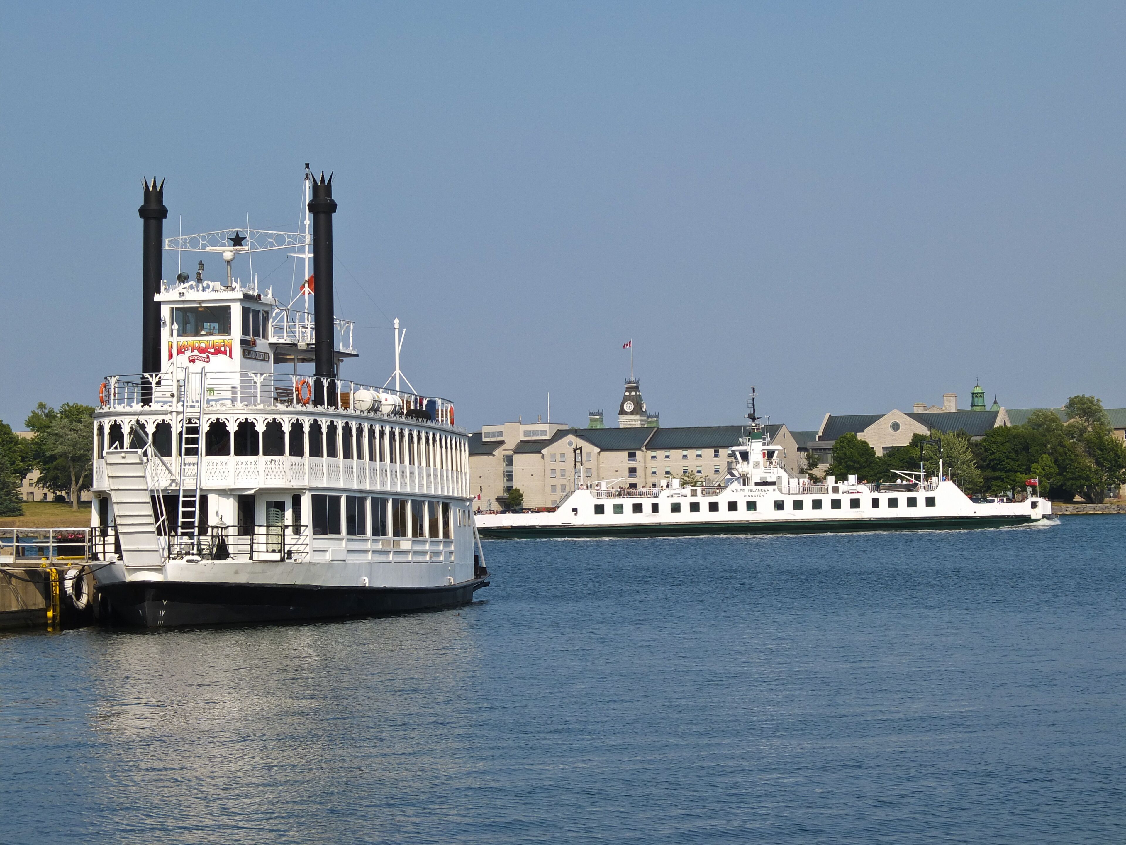 A riverboat with a white exterior and a prominent chimney, docked near a building with a clock tower and a green roof.