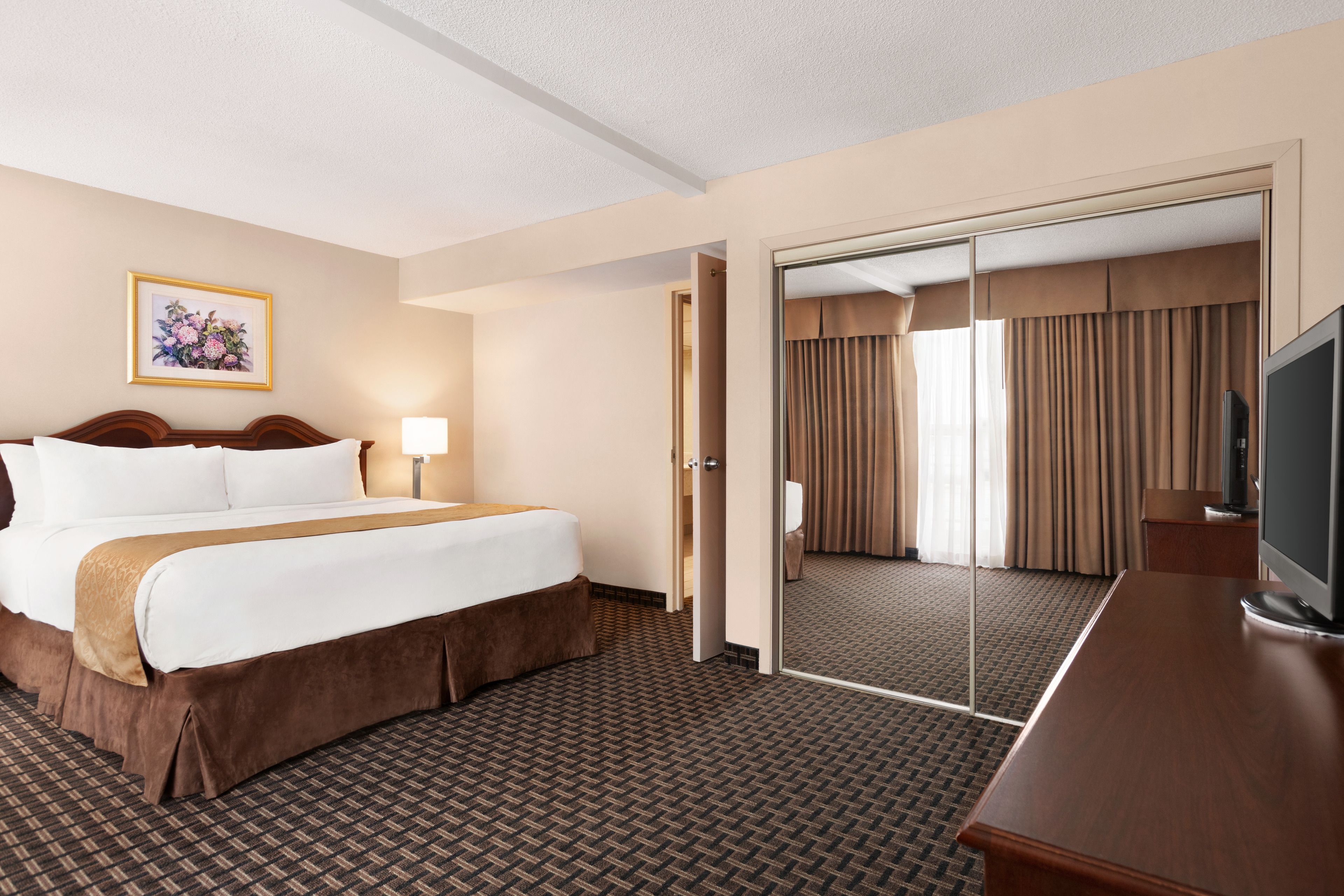 A hotel room with a large bed, a desk with a television, a mirror, and a door leading to a bathroom.