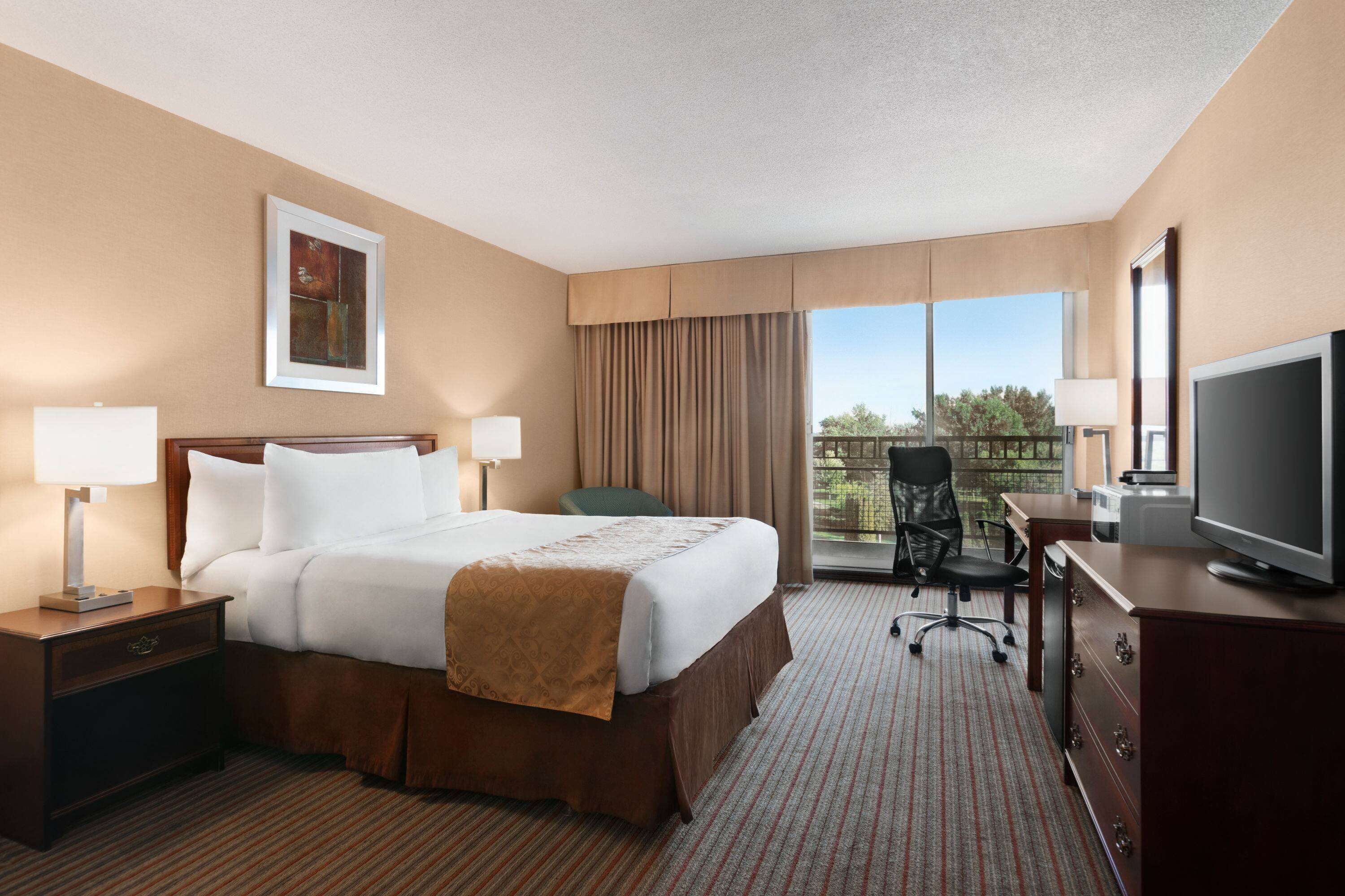 Habitación de hotel con una cama grande, un escritorio con televisor, una silla y una ventana con vistas al exterior.