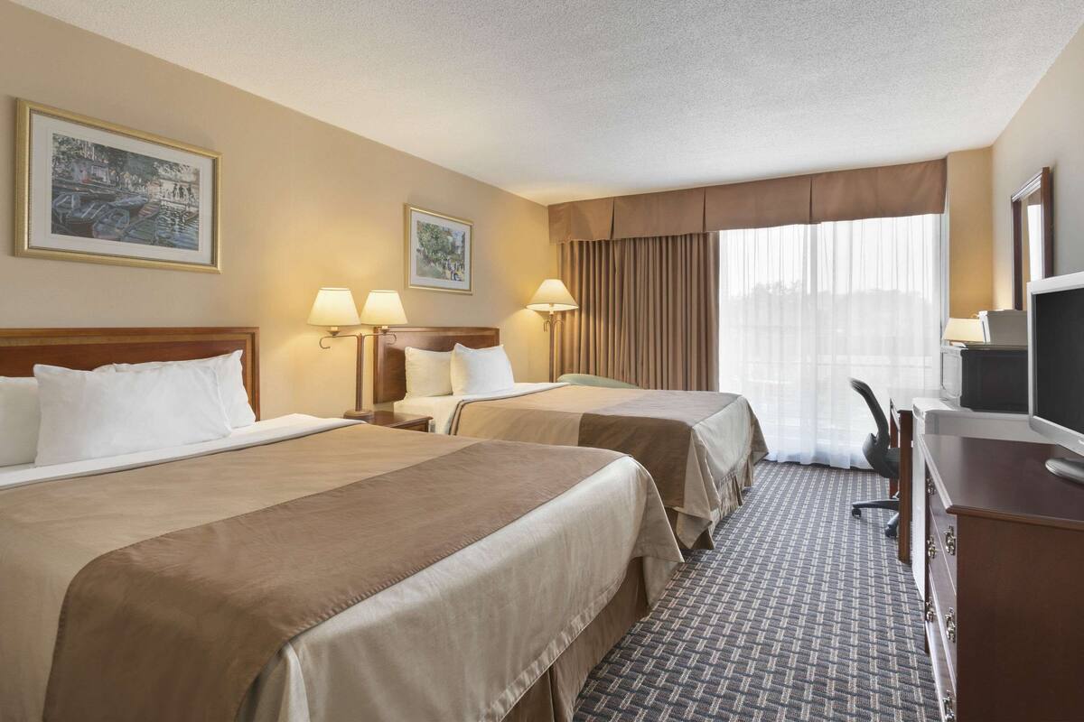 Une chambre d’hôtel avec deux lits, un bureau avec une télévision et une chaise. Il y a deux lampes fixées au mur et une fenêtre avec des rideaux.