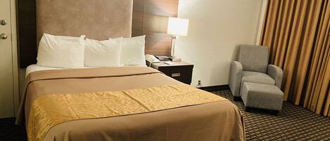 A hotel room with a large bed, a desk with a lamp, a chair, and a window with curtains.