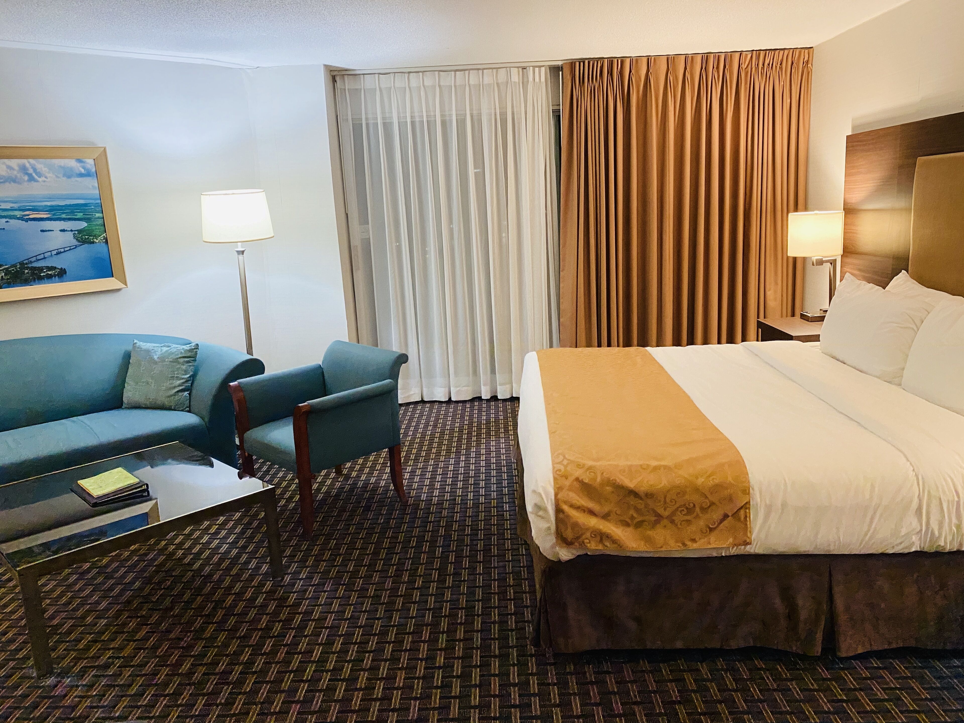A hotel room with a bed, a sofa, a chair, a desk, a lamp, and a painting on the wall.