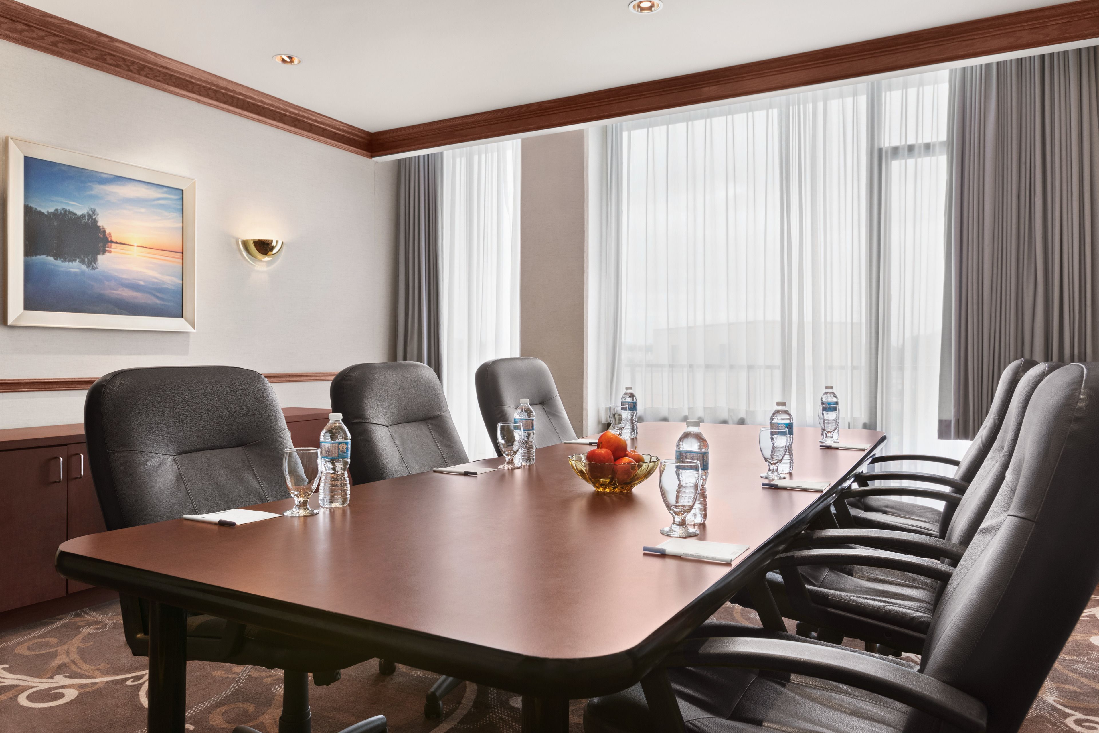 A conference room with a long table, six chairs, water bottles, a fruit bowl, and a painting on the wall.