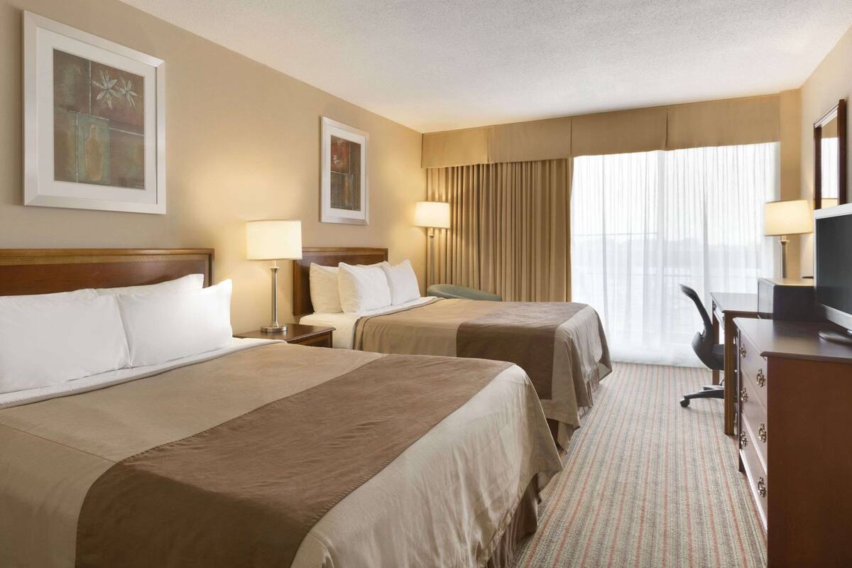 Une chambre d’hôtel avec deux lits, un bureau avec un ordinateur et une grande fenêtre avec des rideaux.