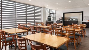 Un vaste espace dédié à la salle à manger, avec de nombreuses tables et chaises en bois, de grandes fenêtres équipées de stores et une cuisine visible à travers ces stores.