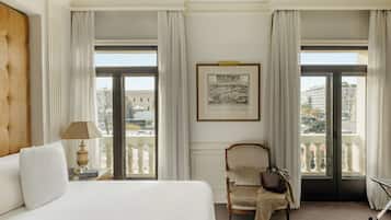 A hotel room with a large bed, a chair with a handbag, a framed picture on the wall, and a balcony with a view of buildings.