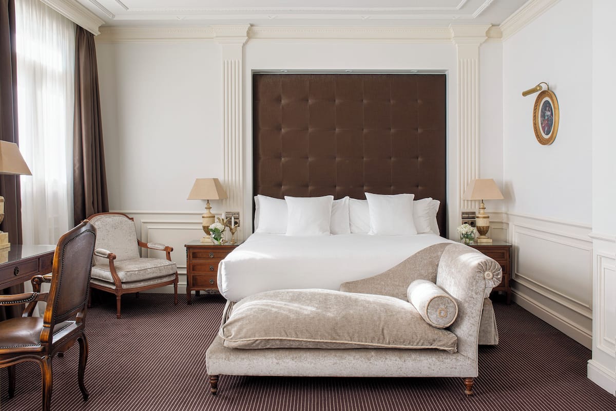 Une chambre d’hôtel dotée d’un grand lit, d’un coin salon avec un fauteuil, d’un bureau et de deux tables de chevet ornées de lampes et d’objets décoratifs.