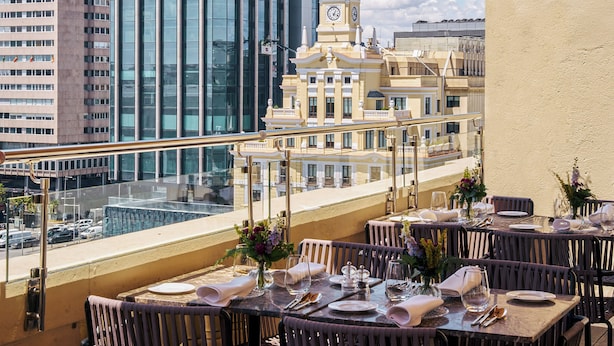 Un restaurante en la azotea con mesas dispuestas para comer, con vistas a un paisaje urbano con edificios modernos y una torre del reloj.