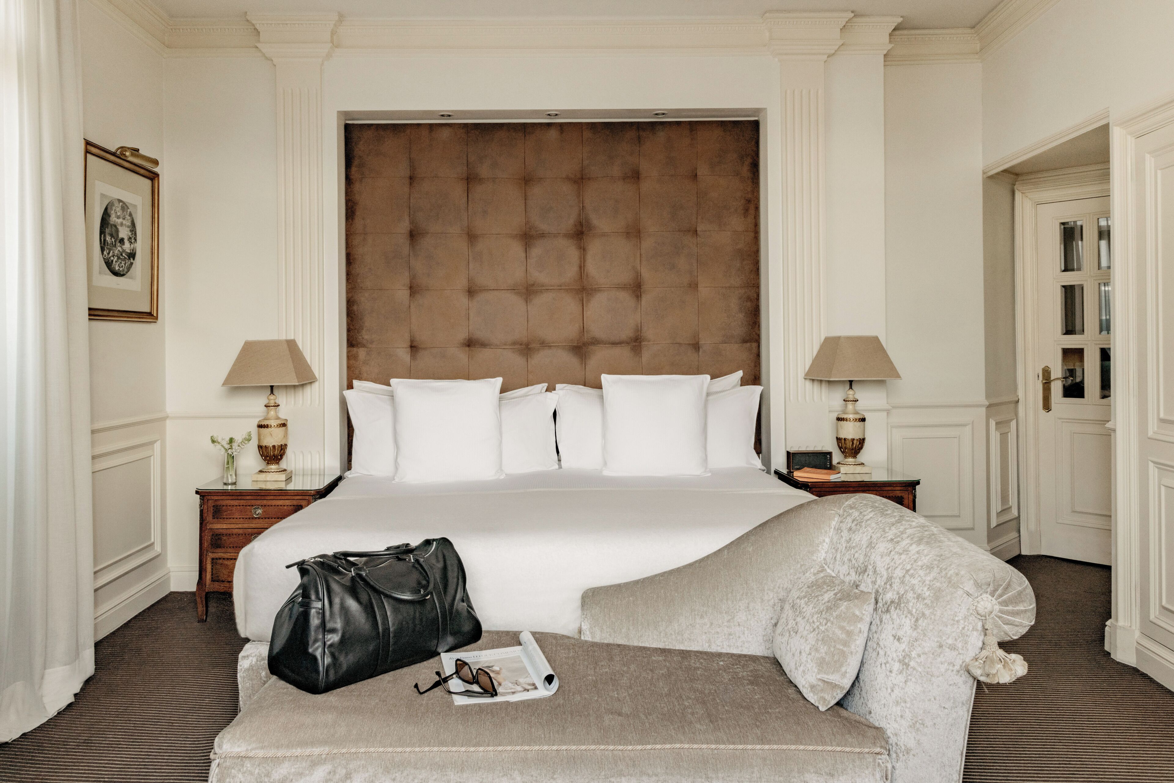 A hotel room with a large bed, two bedside tables, a bench, and a door.