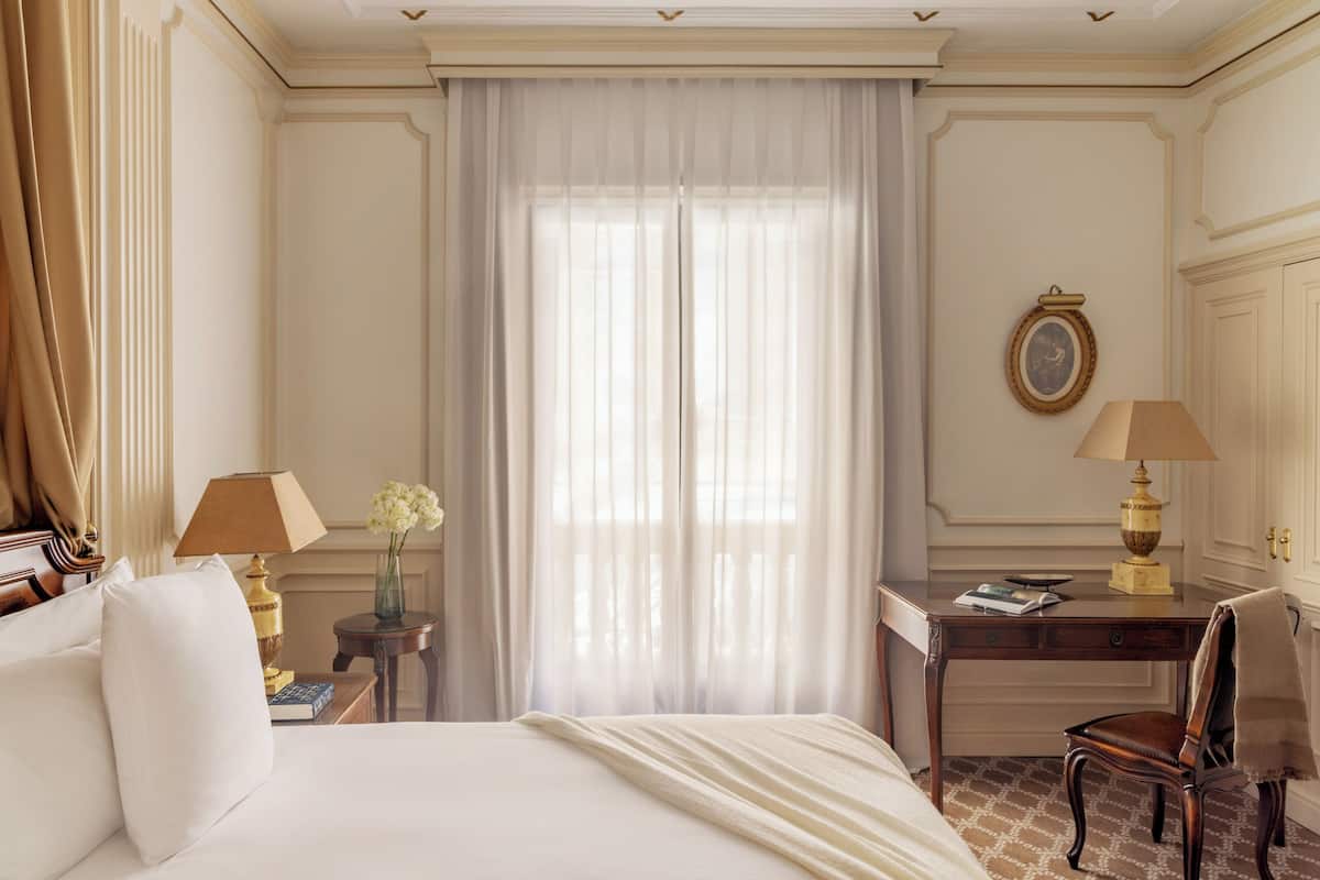 Une chambre d’hôtel comprenant un lit, deux tables de chevet, un bureau, une chaise et une grande fenêtre avec des rideaux légers.