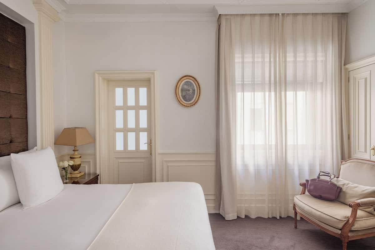 Une chambre d’hôtel avec un grand lit, une chaise, une lampe, une porte et une fenêtre avec des rideaux.